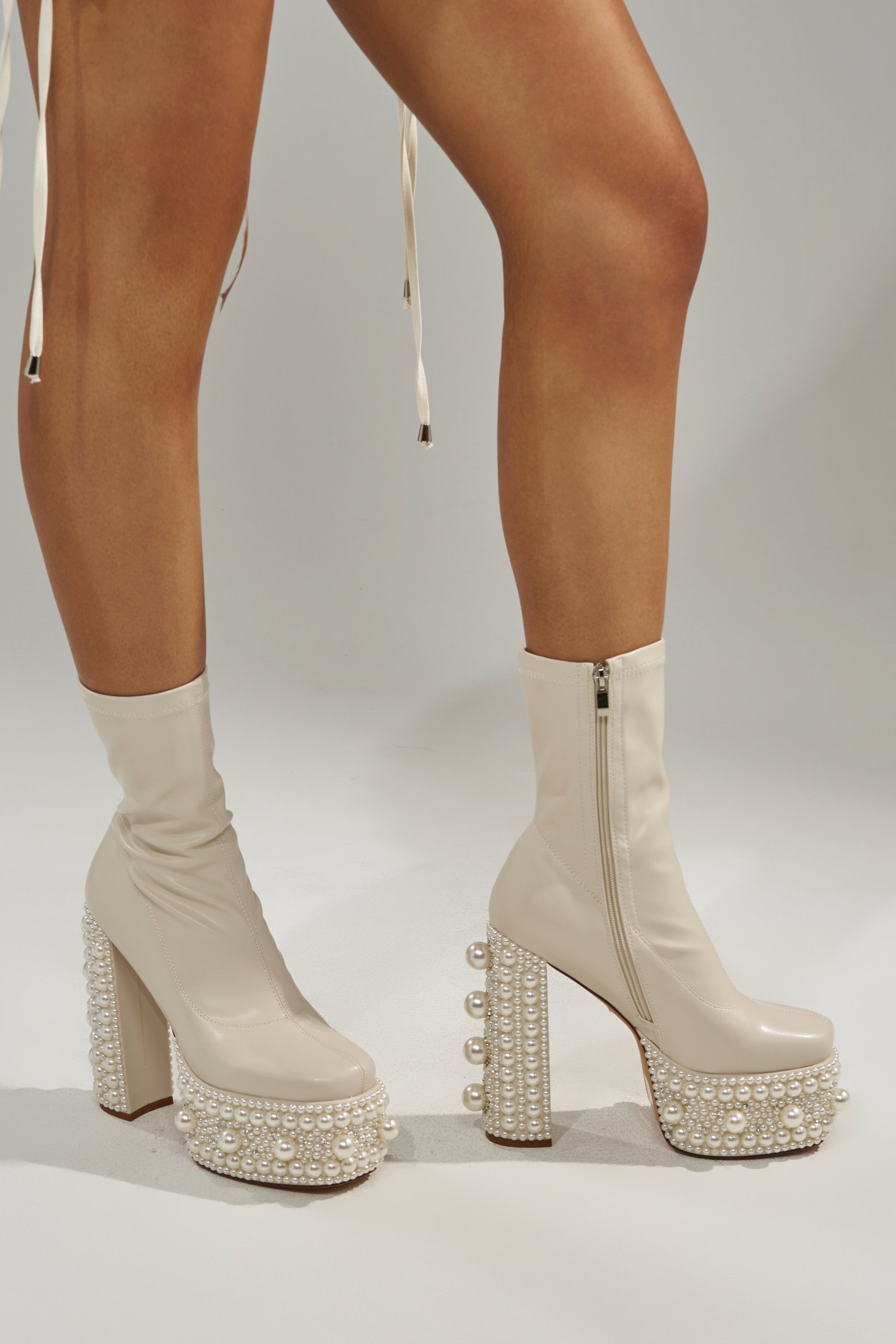 Someone wears the AZALEA WANG ANTARIS BEIGE PEARL DETAIL PLATFORM BOOTIE, featuring chunky high heels and decorative white pearls, while standing against a plain light background. 3