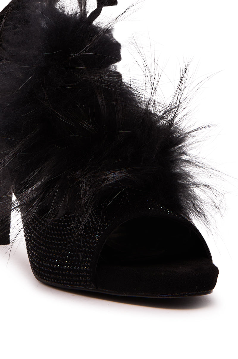 A close-up of the AZALEA WANG ANTIRI BLACK SPARKLE AND FUR SANDAL—a high-heeled, open-toe black sandal with black feathers and rhinestones—set against a white background.