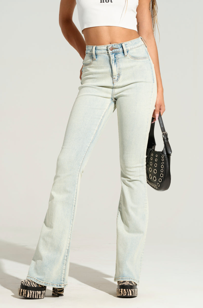 Dressed in the stylish KEEP WISHING DENIM PANT, a person stands confidently wearing light blue, high-rise flare jeans paired with a cropped white top. One hand rests on their hip while the other grips a small black handbag. The chic outfit is completed with platform zebra-print shoes, creating an elegant silhouette against the plain background.