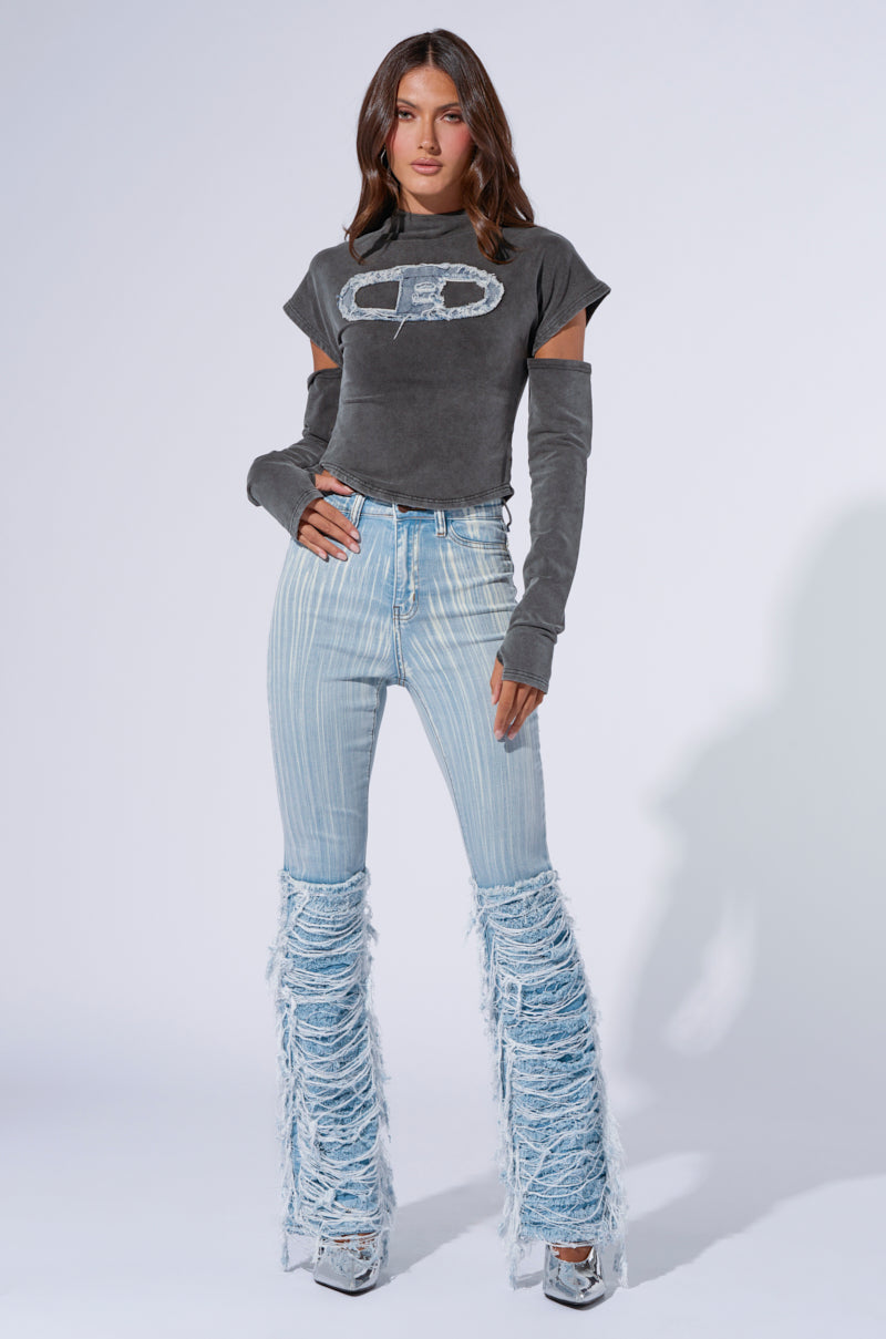 A woman poses against a plain white background, wearing a dark grey cropped t-shirt with cut-out sleeves and a logo on the chest. She pairs it with the STOP AND STARE DENIM PANT, featuring light wash denim high-waisted jeans with distressed detailing from the knees down, and metallic platform shoes.