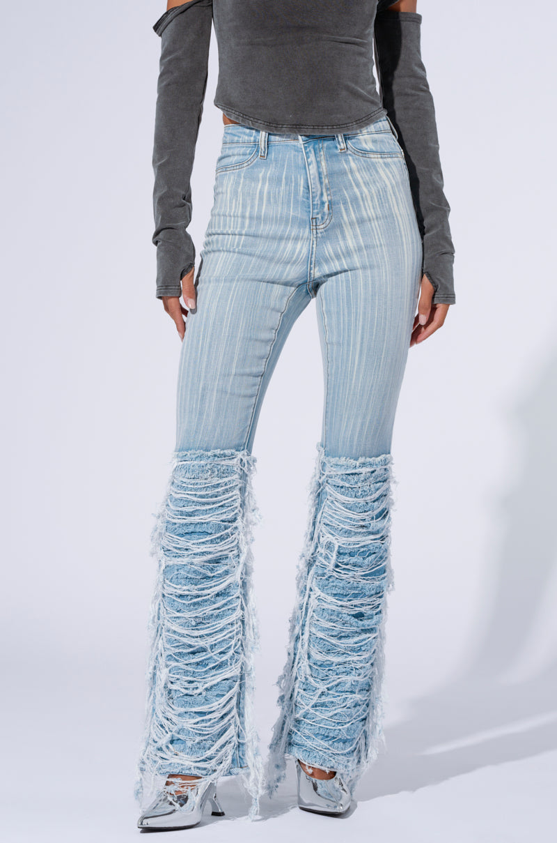 An individual is dressed in a grey, off-the-shoulder long sleeve crop top, complemented by the STOP AND STARE DENIM PANT featuring ultra distressed detailing from the knee down. The light wash denim ensemble is styled with silver pointed-toe shoes against a plain white background.