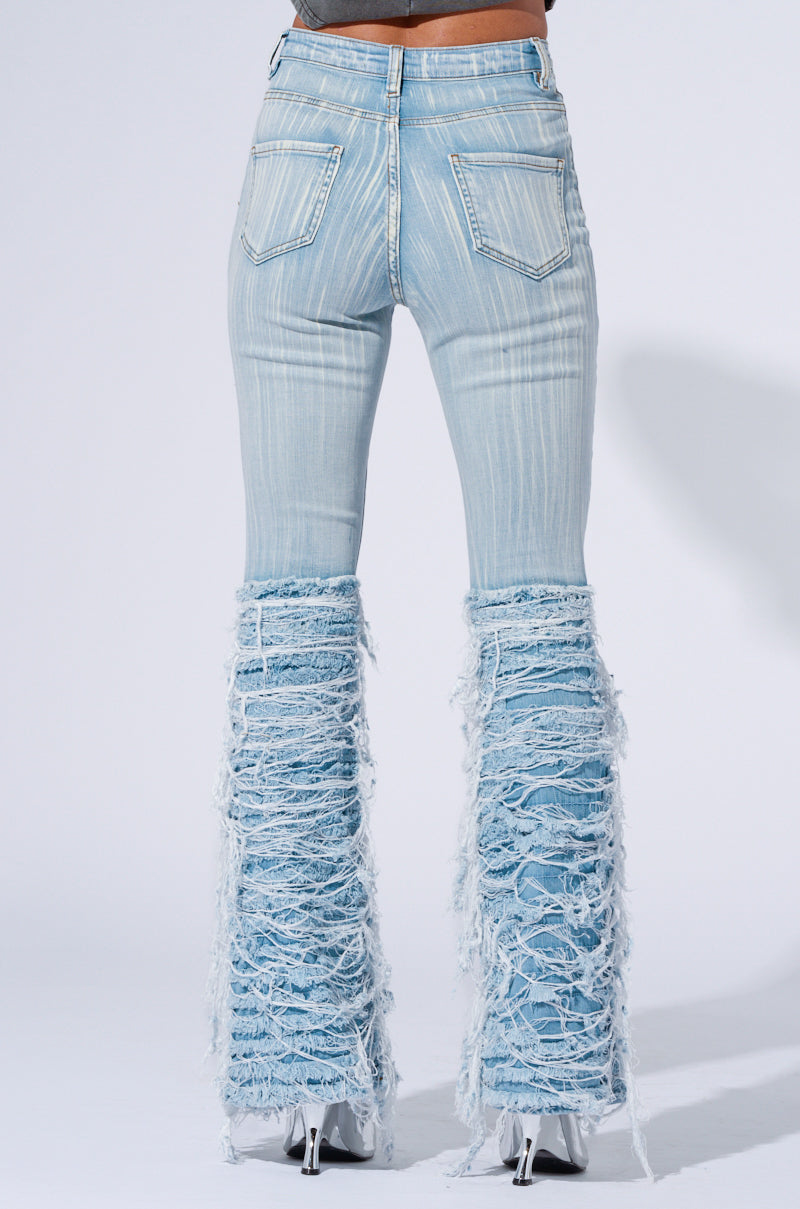 Person wearing the STOP AND STARE DENIM PANT, light blue distressed flare jeans with heavy fraying starting from the knee down. The high-rise design features two visible back pockets, and the person is wearing white high-heeled boots. The background is plain white.