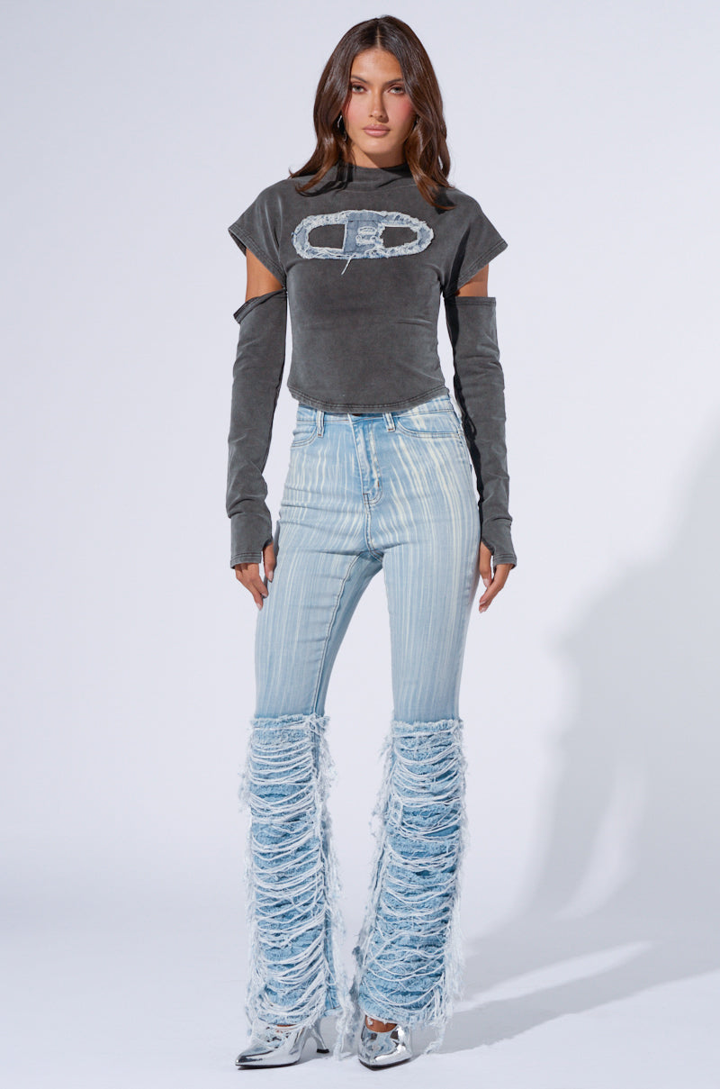 A woman with long brown hair stands against a white background. She is wearing a gray distressed crop top with long sleeves, high-waisted blue and white striped jeans featuring distressed detailing, known as the "STOP AND STARE DENIM PANT," and light blue fringed pants over silver heeled shoes.
