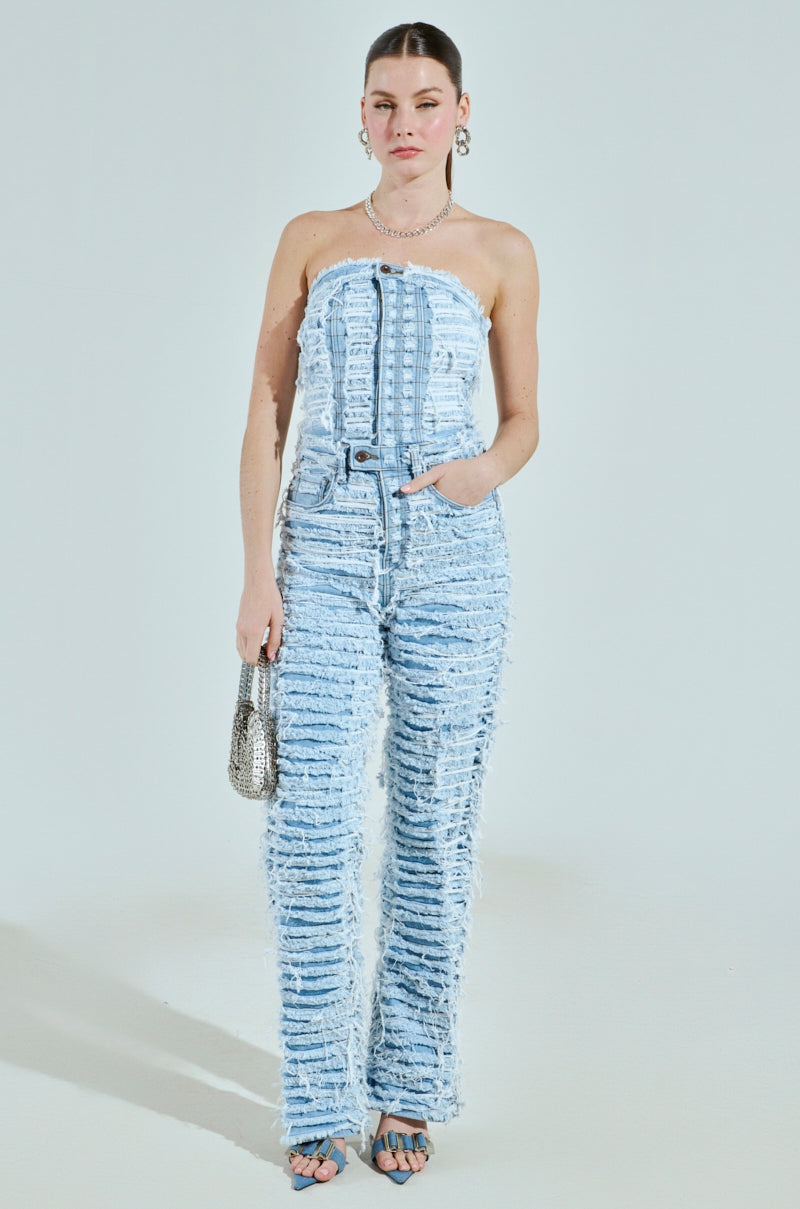 A woman models the "I DON'T CHASE I ATTRACT" distressed light blue denim jumpsuit with frayed edges and princess seams. She holds a small metallic purse, wears hoop earrings and a pearl necklace, and poses with her hair pulled back against a plain background.