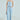 A woman models the "I DON'T CHASE I ATTRACT" distressed light blue denim jumpsuit with frayed edges and princess seams. She holds a small metallic purse, wears hoop earrings and a pearl necklace, and poses with her hair pulled back against a plain background.