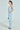 A woman stands in a I DON’T CHASE I ATTRACT distressed light blue denim jumpsuit with Princess Seaming, her hands in the pockets. Her long hair is tied back, and she's wearing hoop earrings. She holds a small beaded handbag and wears high-heeled sandals against a plain background.
