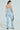 Poised in the "I DONT CHASE I ATTRACT" distressed light blue denim jumpsuit with frayed accents, a woman stands carrying a small sparkly purse, wearing high heels. Her hair is tied back as she faces away from the camera against a plain background.