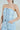 Someone confidently poses in the "I DONT CHASE I ATTRACT" distressed light blue denim jumpsuit with frayed edges and layered patterns. Styled elegantly with a silver chain necklace and hoop earrings, they casually rest one hand in their pocket against a plain backdrop.
