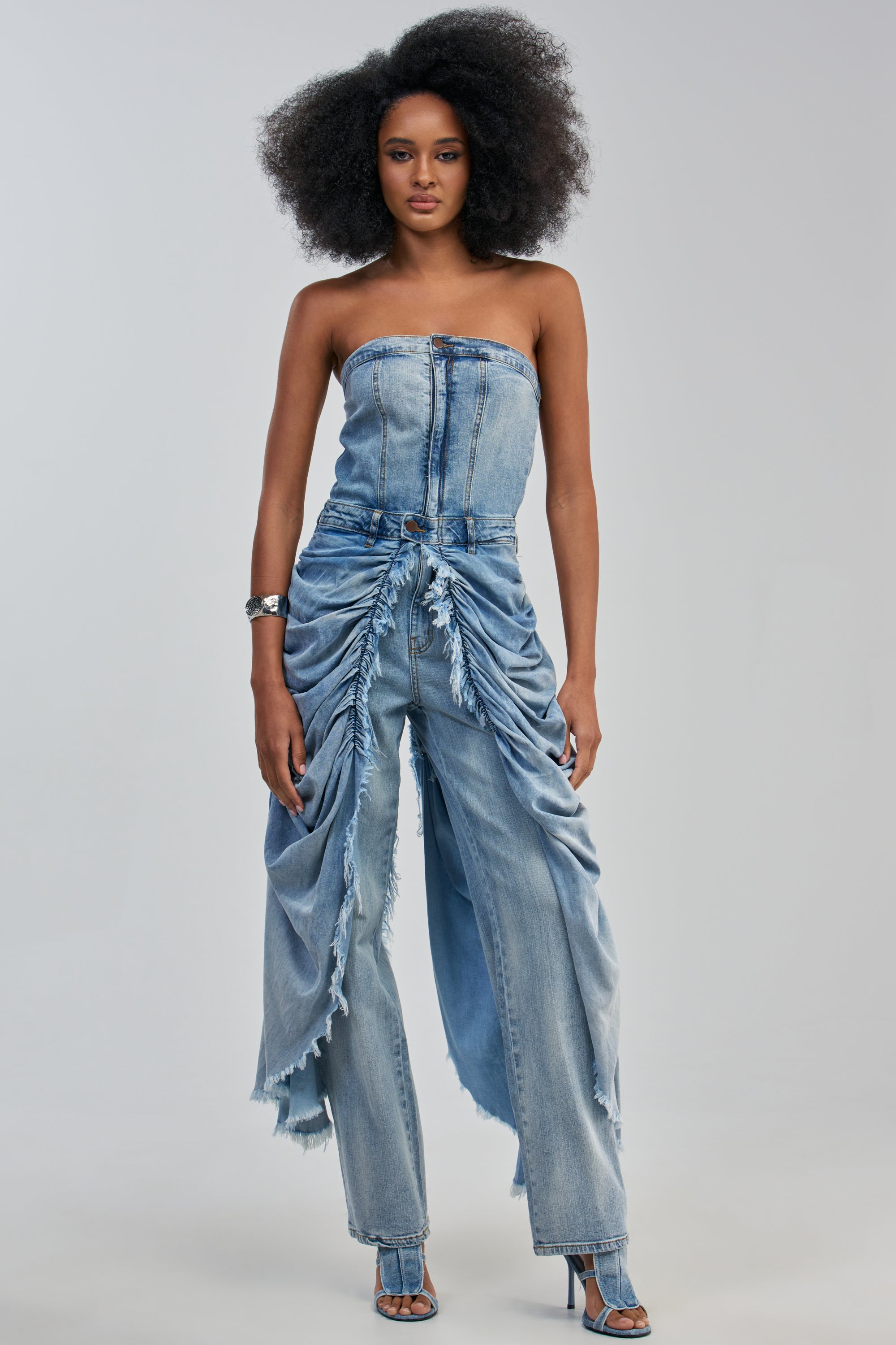 Wearing the REBEL HEART STRAPLESS DENIM JUMPSUIT—featuring high-waisted, ruched, and frayed pants with layered panels—a woman with natural hair stands confidently in heeled sandals against a plain background, embodying modern streetwear jumpsuit style.