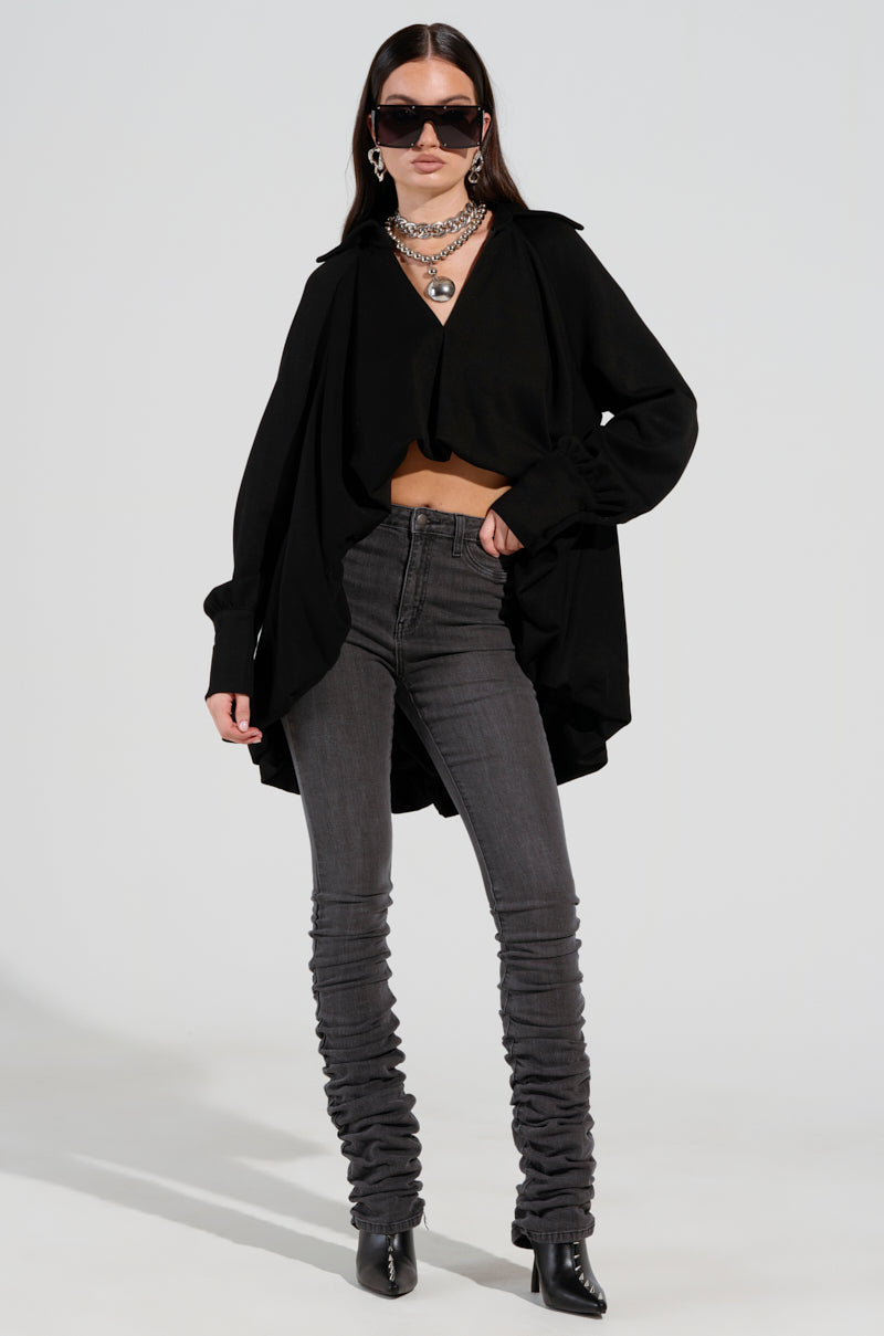 A woman confidently poses in the STACKS ON STACKS DENIM PANT, paired with a black draped top and dark sunglasses. She accessorizes her look with layered silver necklaces and black ankle boots, standing against a plain background and exuding effortless style. 1