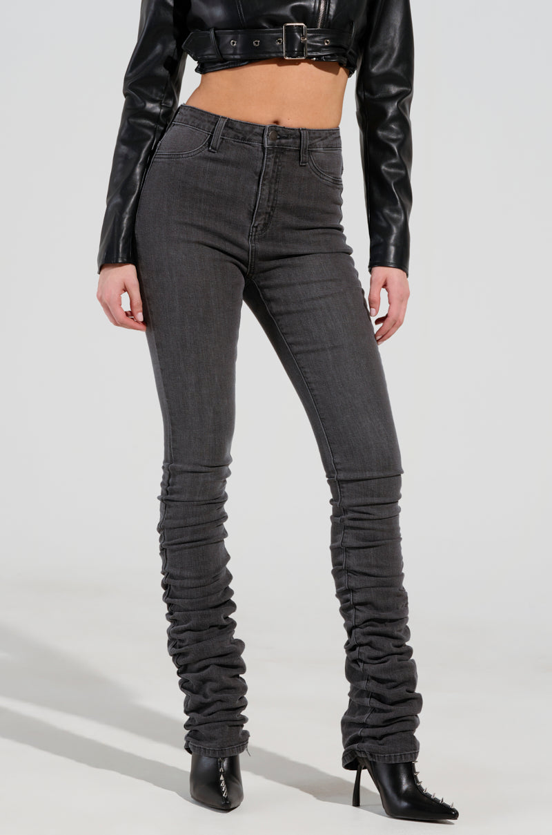 A person wearing the STACKS ON STACKS DENIM PANT, which features a high-waisted fit and dark gray wash with a ruched design from the knee down, paired with a black cropped leather jacket. They complete their chic look with sophisticated heeled black ankle boots. 2