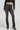 A person wearing the STACKS ON STACKS DENIM PANT, which features a high-waisted fit and dark gray wash with a ruched design from the knee down, paired with a black cropped leather jacket. They complete their chic look with sophisticated heeled black ankle boots.