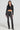 Wearing a black cropped leather jacket, layered necklaces, the STACKS ON STACKS DENIM PANT in a dark high-rise skinny leg style, and black heeled boots, the person stands confidently against a plain background. The ensemble exudes a stylish and edgy vibe.
