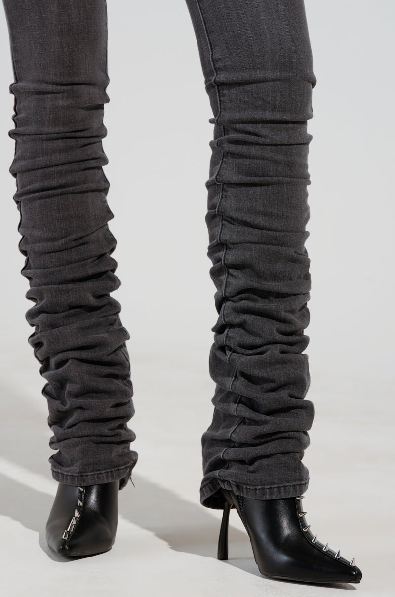 A person wearing STACKS ON STACKS DENIM PANTS and black high-heeled boots with metal studs on the toes stands on a light surface. 7