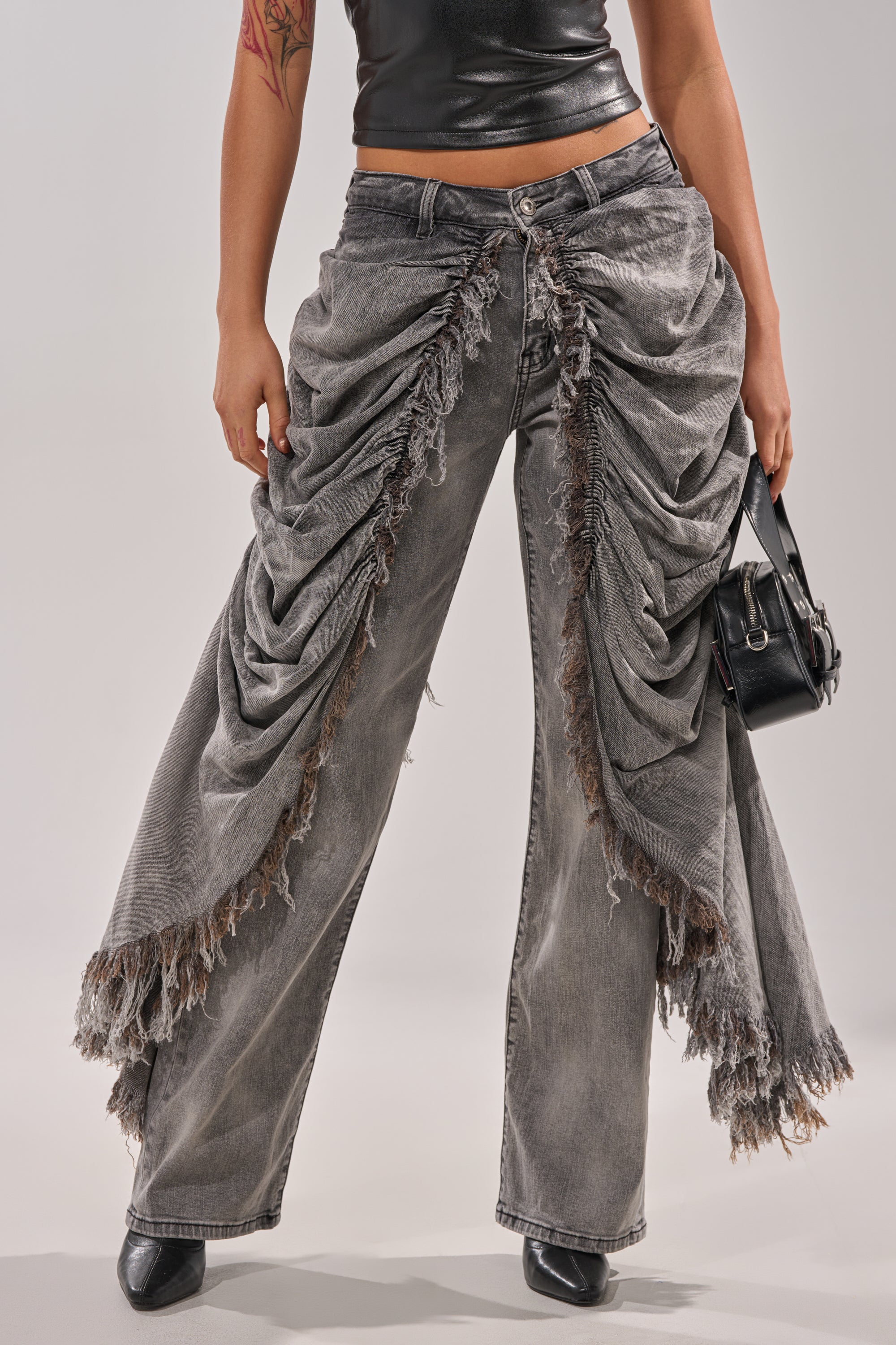 A person showcases avant-garde street style in WOW WOW WOW DENIM PANT—grey high-rise flares with ruched, frayed side panels—paired with a black faux leather top, black handbag, and pointed shoes. Only their lower torso and legs are visible.