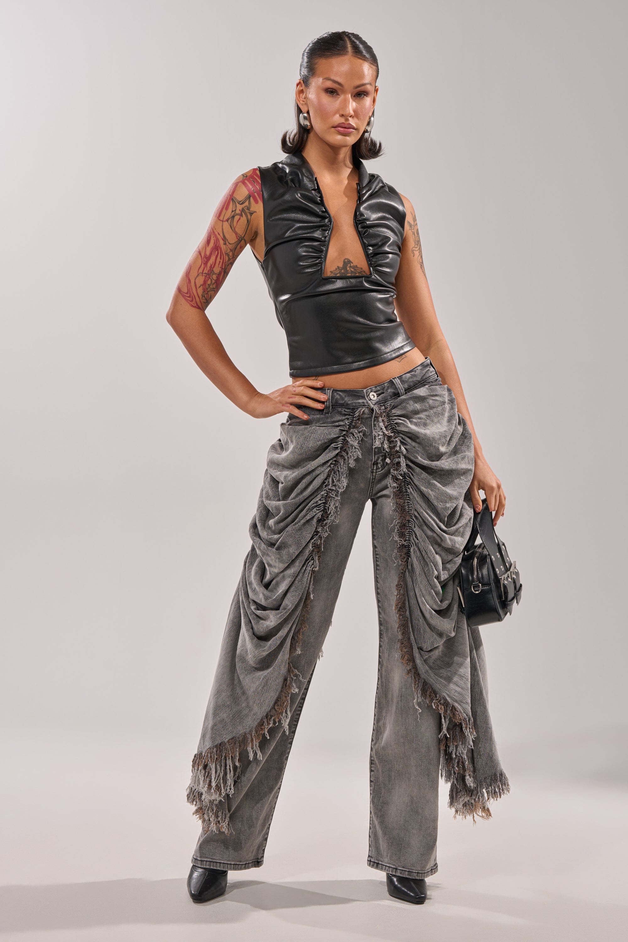 A woman poses confidently in the WOW WOW WOW DENIM PANT with ruched details and frayed hems, styled with a black leather sleeveless top, pointed shoes, and a sleek black handbag. Her wavy hair completes the chic streetwear look. 1