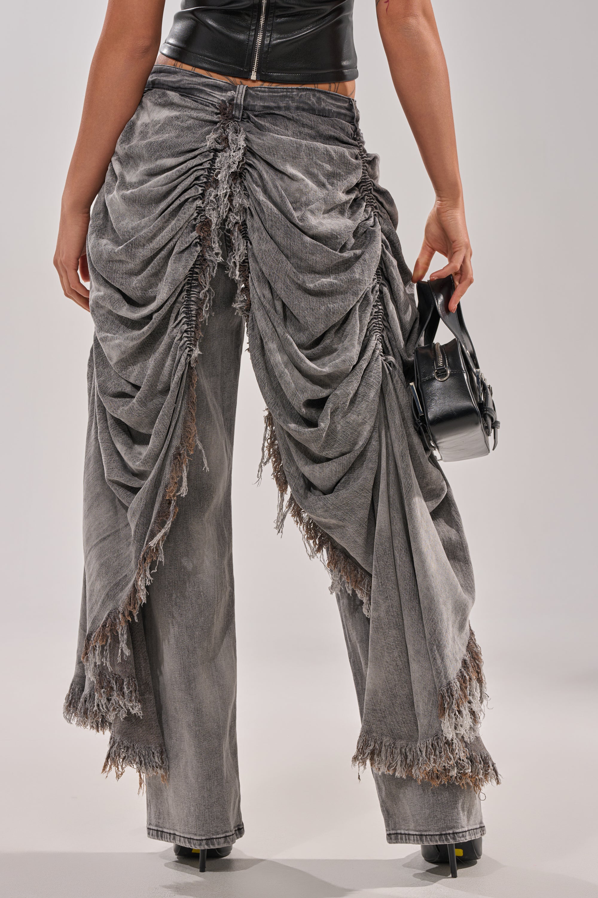 A person wearing WOW WOW WOW DENIM PANT in distressed gray with frayed edges and a black leather top, holding a small black handbag. The photo is taken from behind, highlighting the pants' unique gathered streetwear design. 4