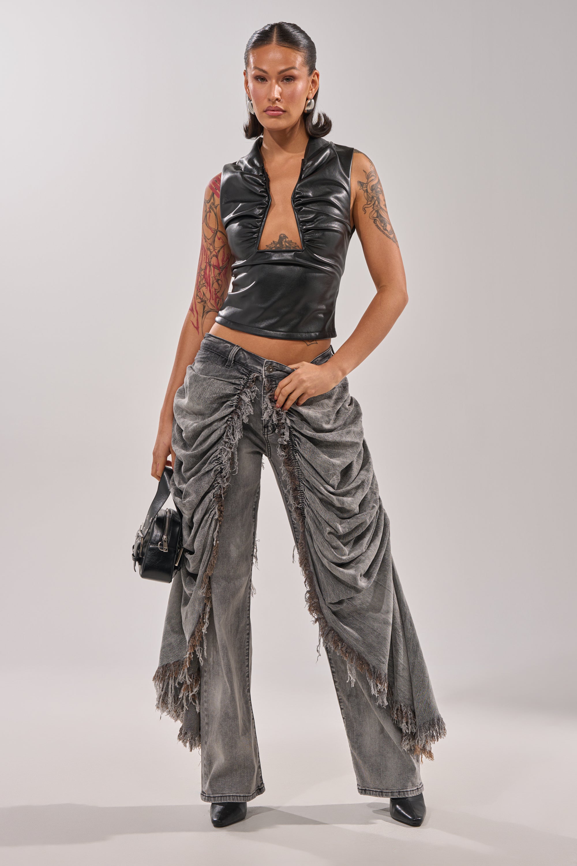 A person stands confidently in a black sleeveless vinyl top and WOW WOW WOW DENIM PANT, styled with dramatic ruching and frayed details. They hold a small black handbag, wear black heels, and display tattoos on one arm. 5