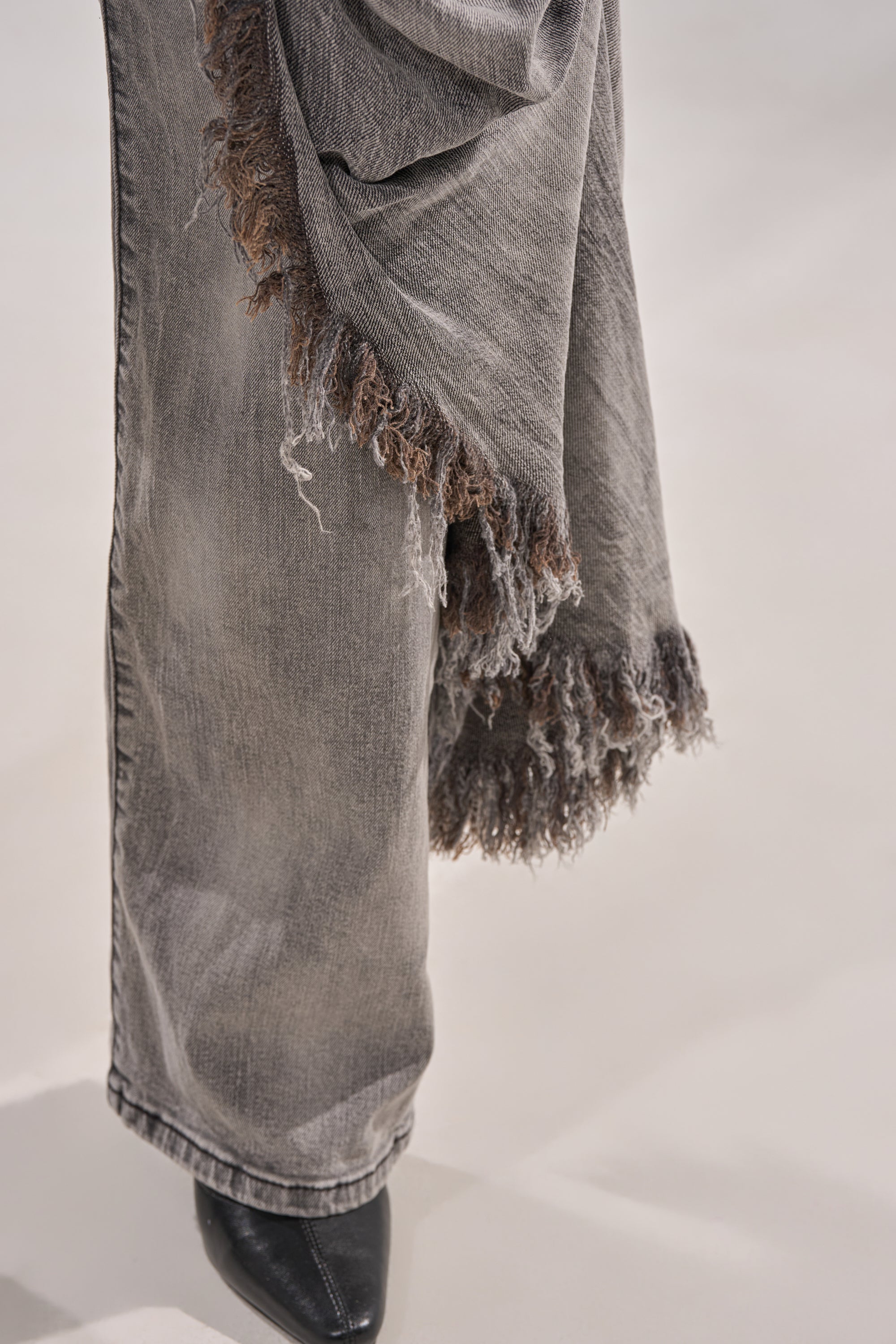 A close-up of a person wearing the WOW WOW WOW DENIM PANT, a high-rise faded grey flare jean with frayed edges, paired with black pointed-toe shoes on a light-colored surface. 7