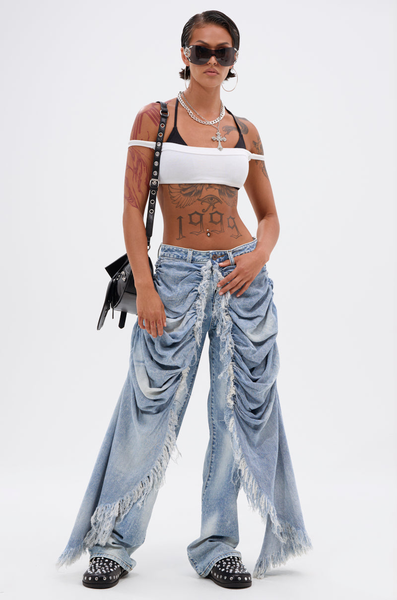 A woman confidently poses against a white background wearing sunglasses, a white tube top, ALL OVER THE PLACE DENIM PANT, studded black shoes, layered necklaces, and a black shoulder bag.