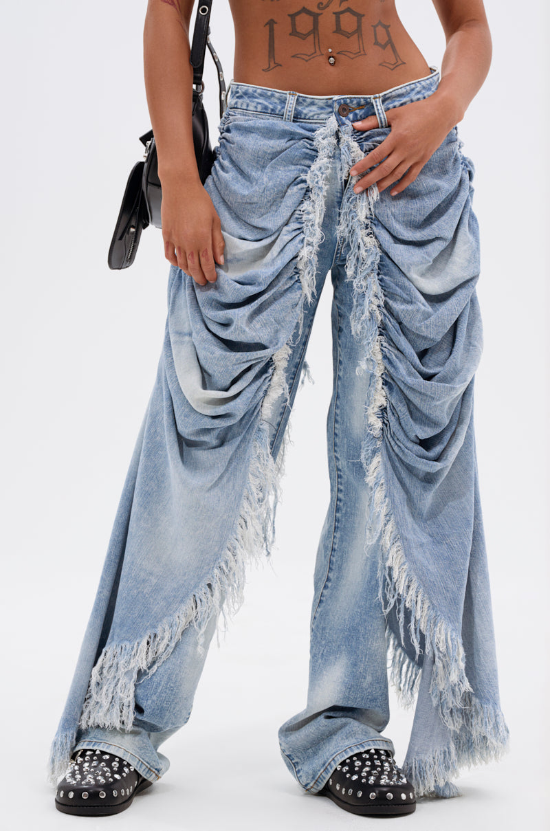 A person stands against a plain white background, wearing the ALL OVER THE PLACE DENIM PANT in light blue with heavy ruching and frayed details, a visible "1999" torso tattoo, a black shoulder bag, and black studded shoes.