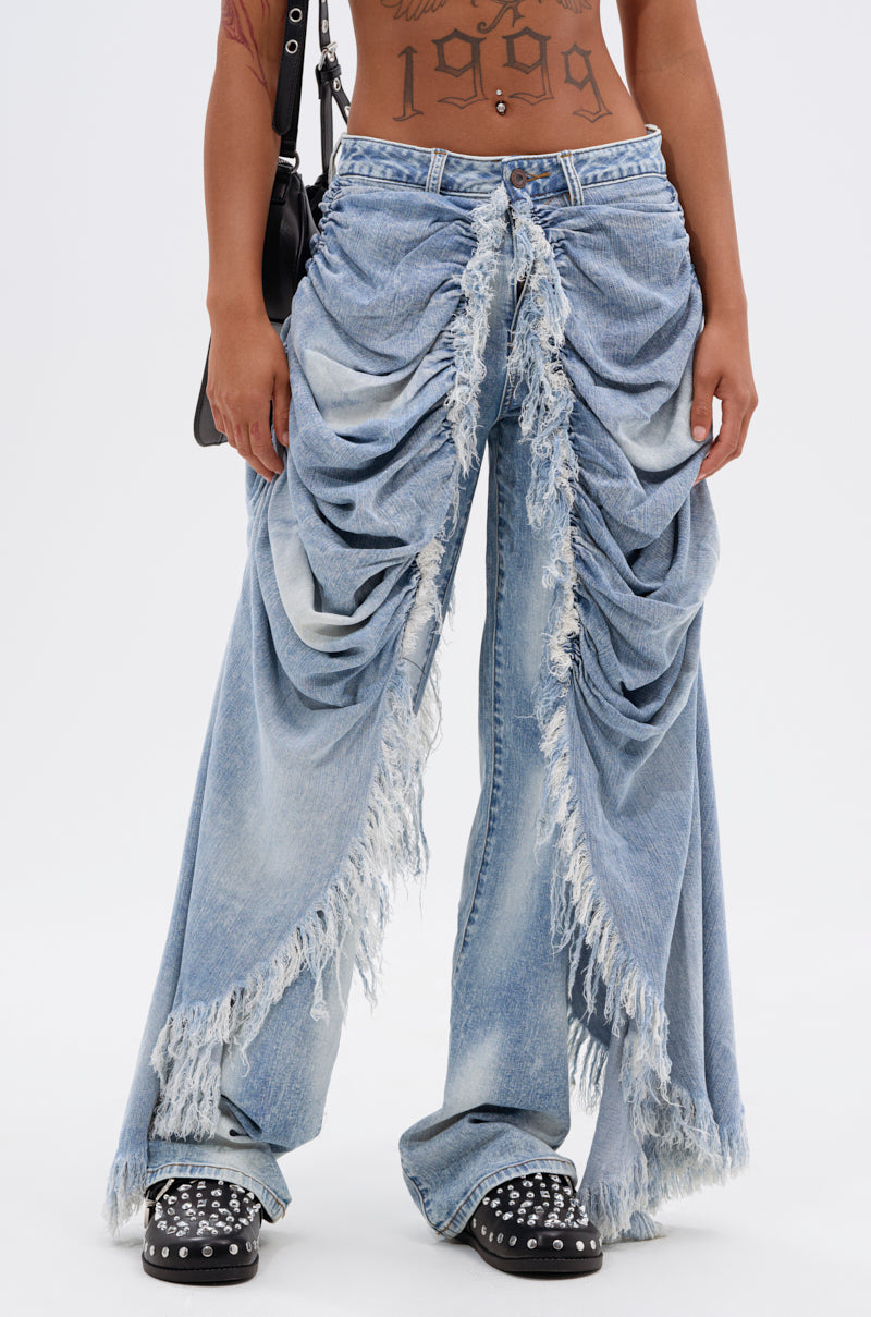 A person wearing ALL OVER THE PLACE DENIM PANT—light blue, acid wash, highly distressed wide-leg jeans with dramatic ruched and frayed details—paired with black studded shoes, a black shoulder bag, and visible arm and torso tattoos.