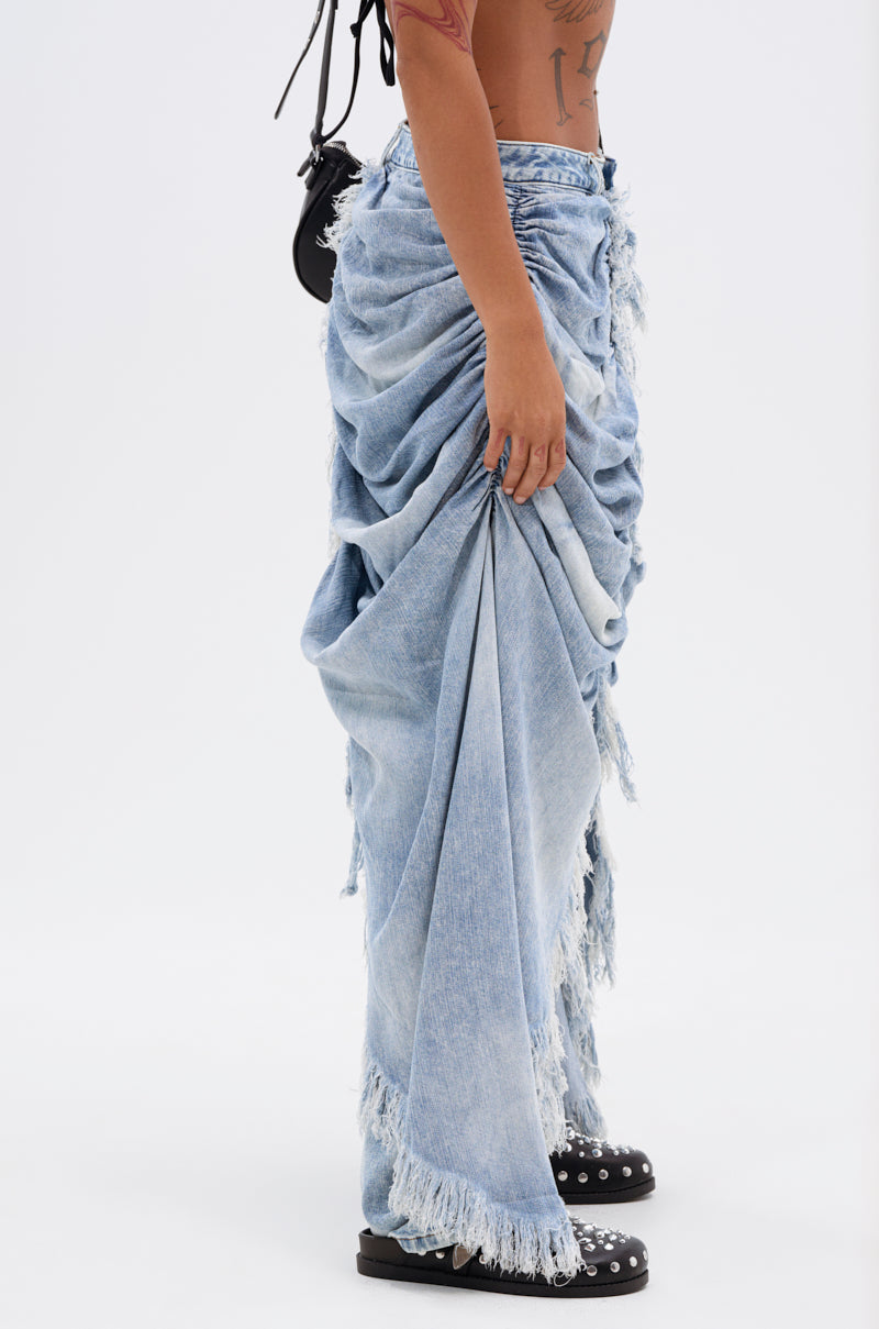A person wears the ALL OVER THE PLACE DENIM PANT, a light blue, distressed denim pant with frayed edges and acid wash effect, paired with black clogs. Only their torso and lower body are visible against a plain white background.