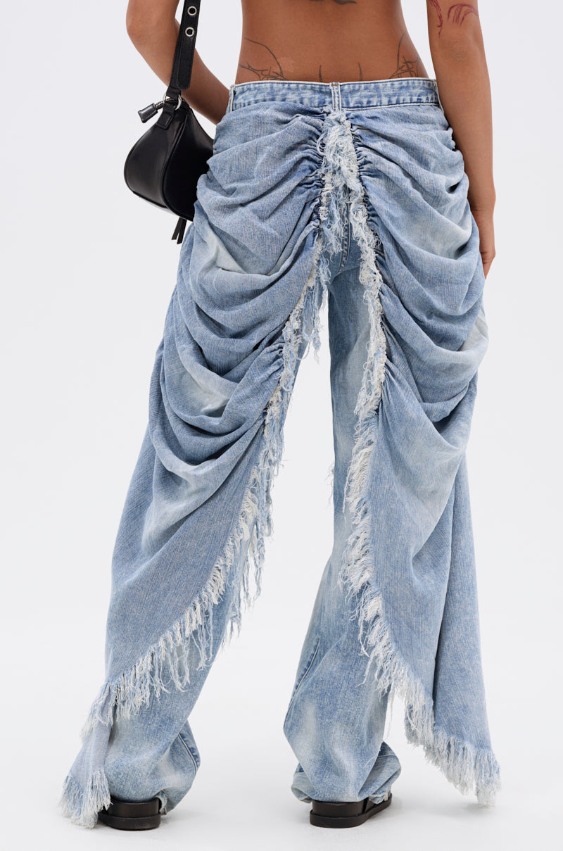 Someone is shown from behind against a plain background, wearing the ALL OVER THE PLACE DENIM PANT in light blue with heavy ruching and frayed back edges, paired with black shoes and a black handbag.