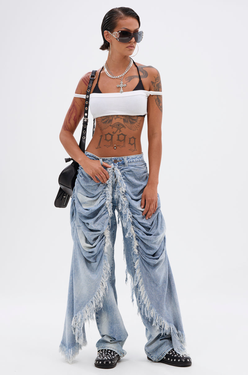 Wearing a white off-shoulder crop top, ALL OVER THE PLACE DENIM PANT in light blue acid wash, dark studded shoes, black shoulder bag, layered necklaces, sunglasses, and showing tattoos on their arms and torso.