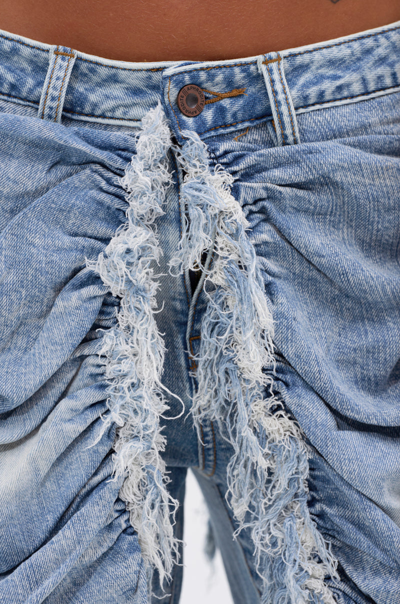 Close-up of the ALL OVER THE PLACE DENIM PANT in light blue, high-rise style with distressed, frayed ruffles by the zipper. Features a fastened metal button and subtle faded detailing on acid wash denim for a textured look.