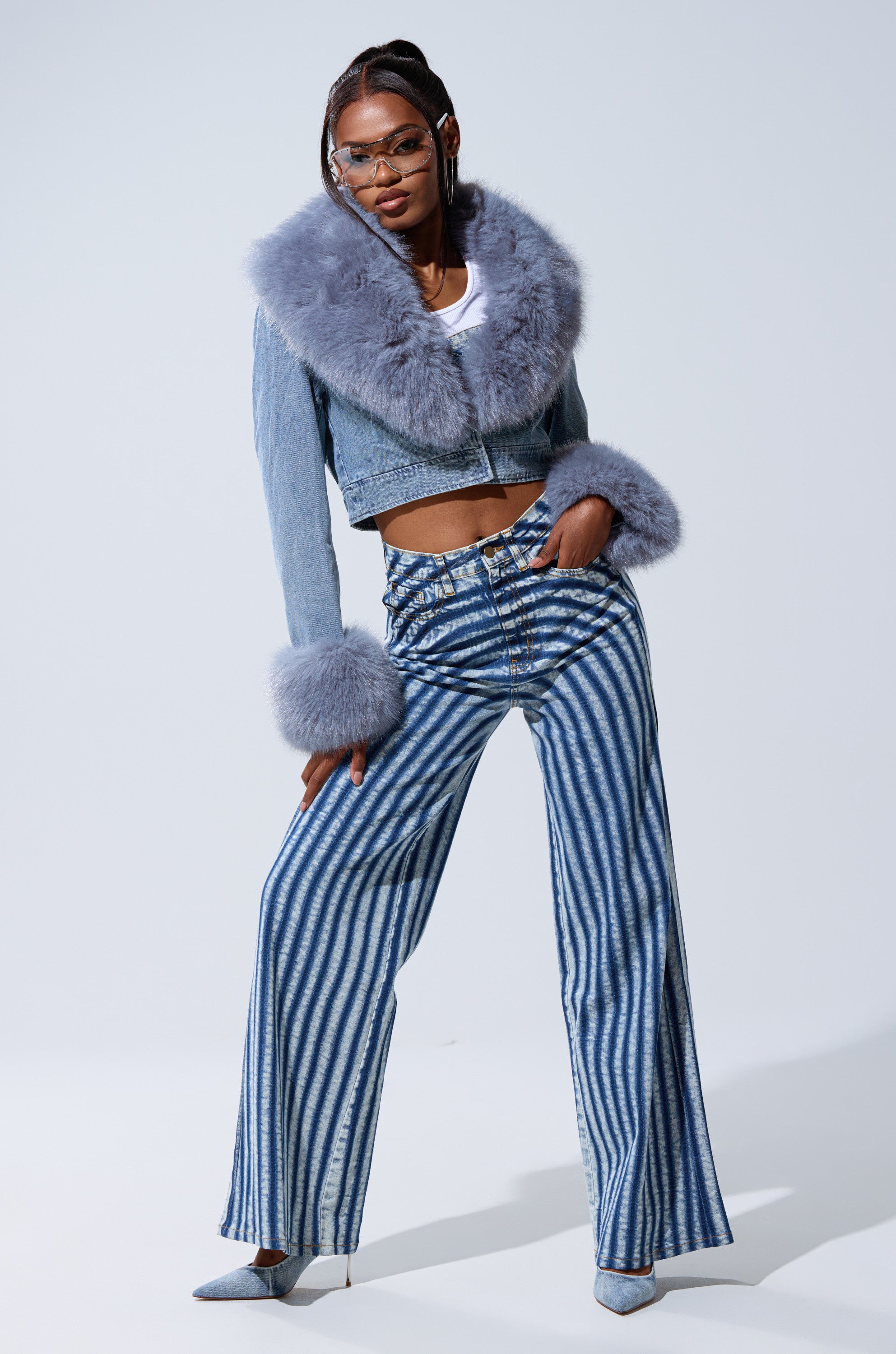 A stylish woman showcases the LETS GET CRAZY STRIPE WIDE LEG DENIM with a cropped denim jacket featuring large gray fur trim, white top, pointed heels, and clear glasses as she poses confidently against a plain light background.