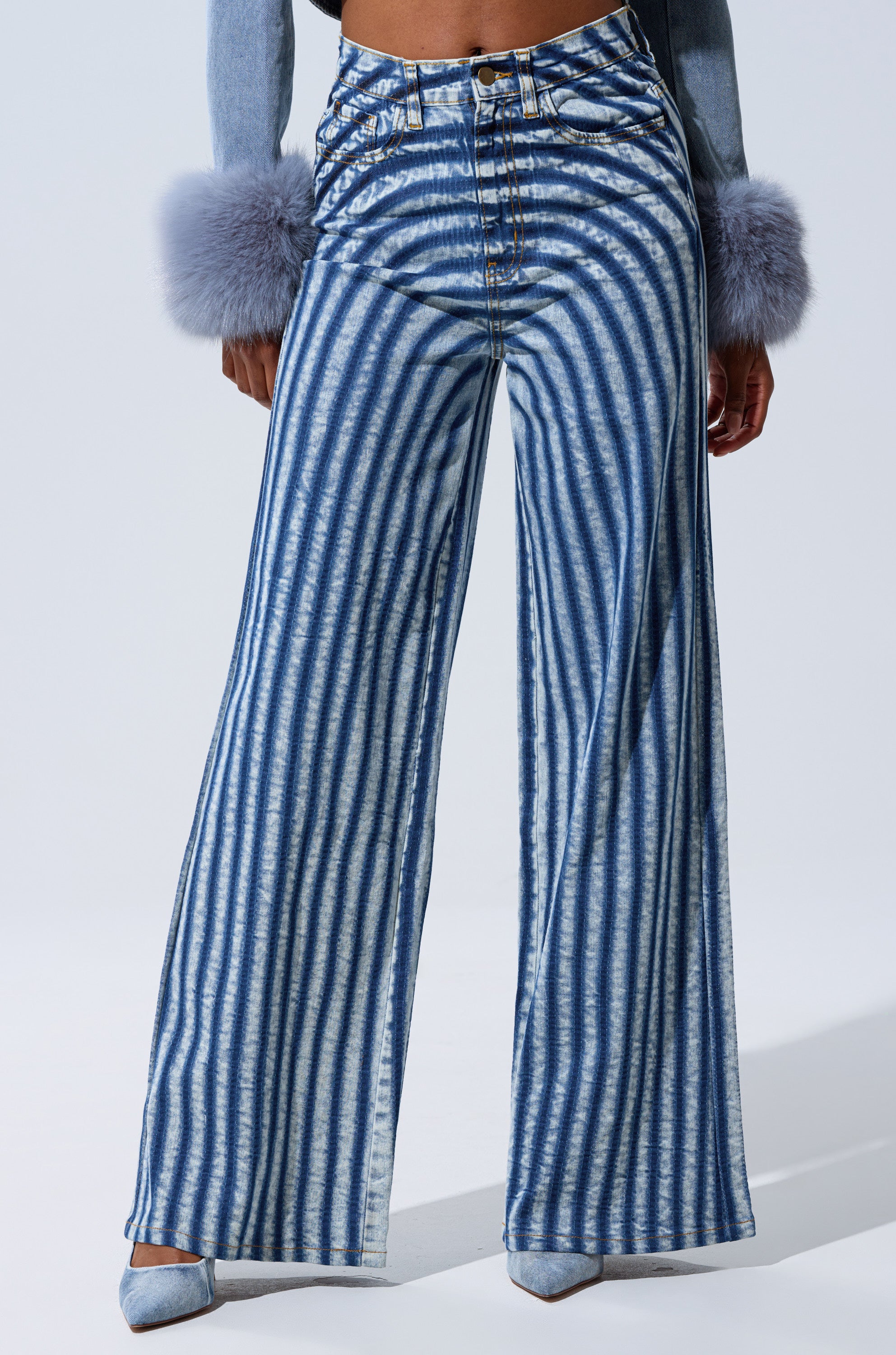 A person wearing LETS GET CRAZY STRIPE WIDE LEG DENIM, paired with light blue pointed-toe heels and a top with fluffy gray cuffs. The image is cropped from the waist down.