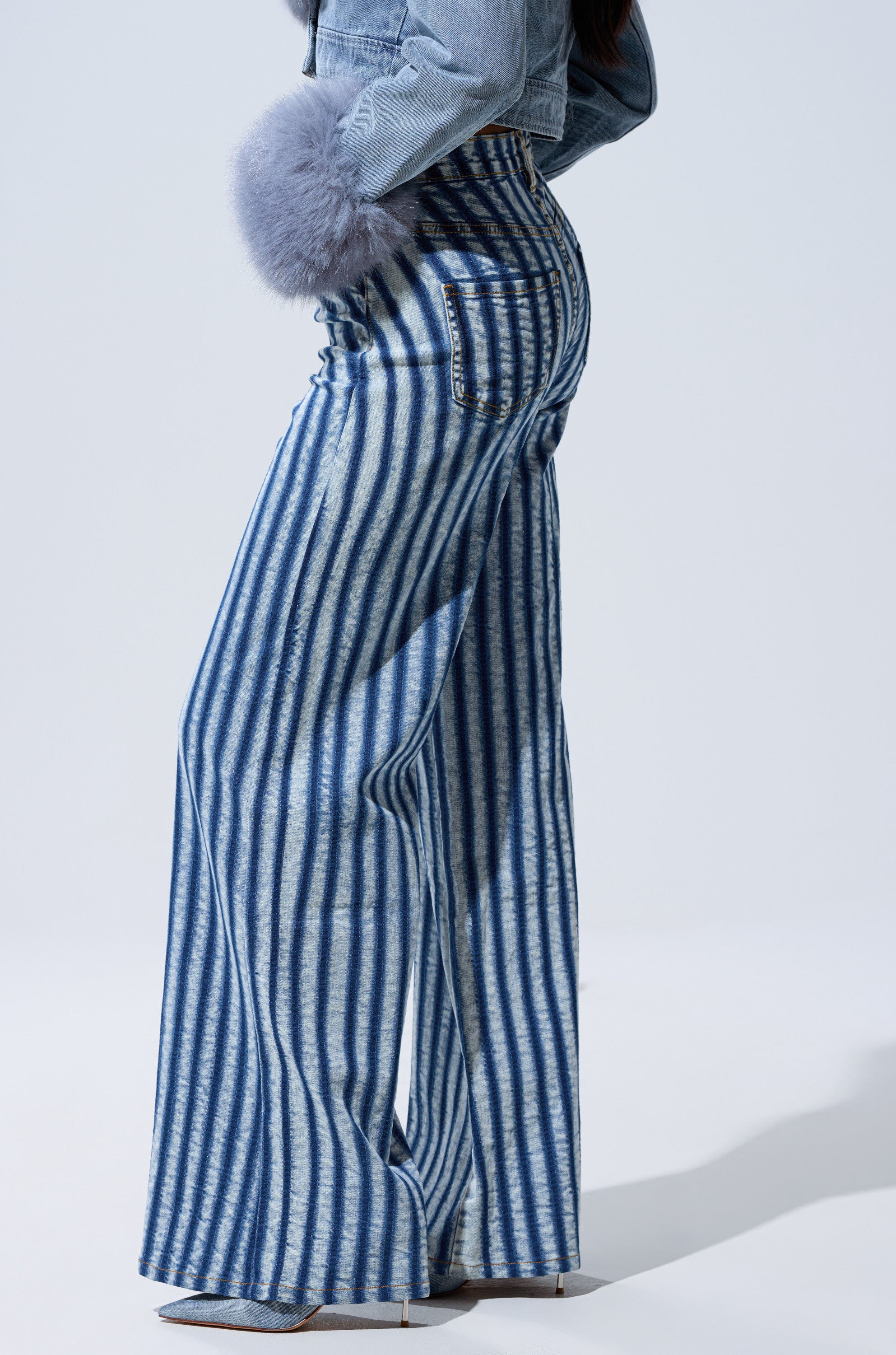 A person wears the LETS GET CRAZY STRIPE WIDE LEG DENIM jeans with a light blue jacket featuring fluffy gray cuffs and pointed shoes, standing on a plain light background.