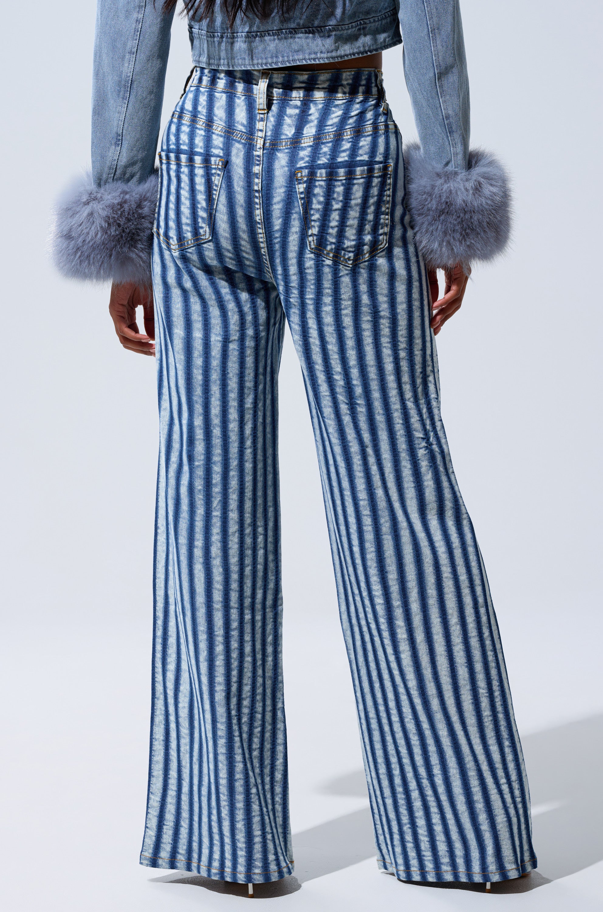A person shown from the back wears the LETS GET CRAZY STRIPE WIDE LEG DENIM in high-rise blue and white stripes, paired with a cropped denim jacket and cuffs featuring large, fluffy gray fur trim.