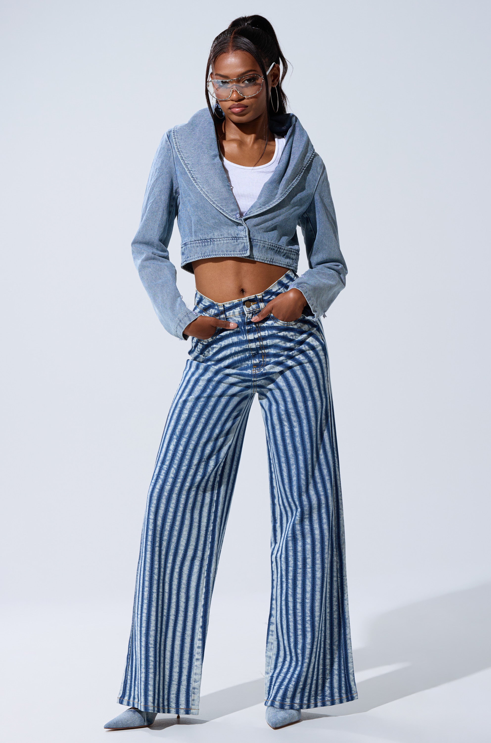 A woman stands confidently with her hands in her pockets, wearing the LETS GET CRAZY STRIPE WIDE LEG DENIM jeans, a cropped denim jacket, white crop top, pointed shoes, and sunglasses against a plain background.