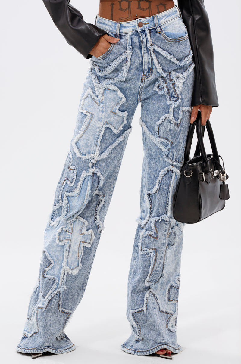 Wearing the CROSSED OUT DENIM DISTRESSED PANT in light blue with large frayed cross patterns, styled with a black leather jacket and holding a black handbag. Only the lower torso and wide-leg silhouette are visible. 0