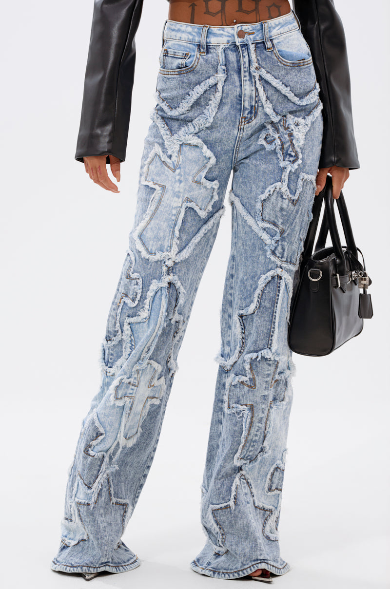 A person wearing the CROSSED OUT DENIM DISTRESSED PANT—light blue, wide-leg jeans with large, frayed cross patterns—paired with a black leather jacket and small black handbag, shown from shoulders to feet. 1