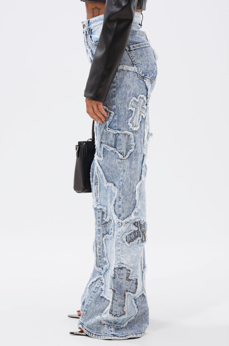 A person stands sideways in the CROSSED OUT DENIM DISTRESSED PANT, light blue high-rise jeans with large cross patches, styled with a black leather long-sleeve top, open-toe heels, and a purse against a plain white background. 2