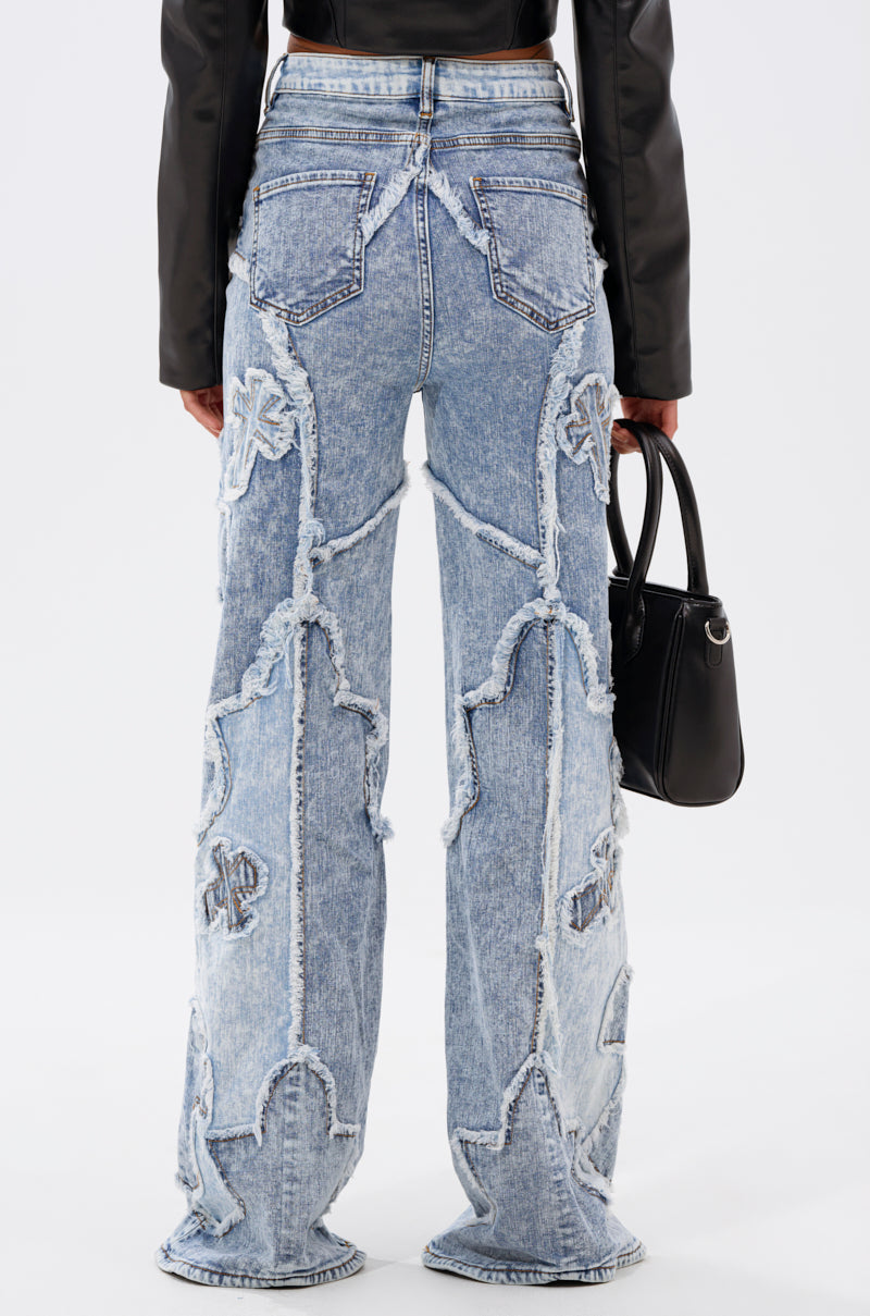 Person seen from the back wearing the CROSSED OUT DENIM DISTRESSED PANT in light blue—high-rise, wide-leg jeans with frayed applique designs—paired with a black long-sleeve top and holding a small black handbag. 3