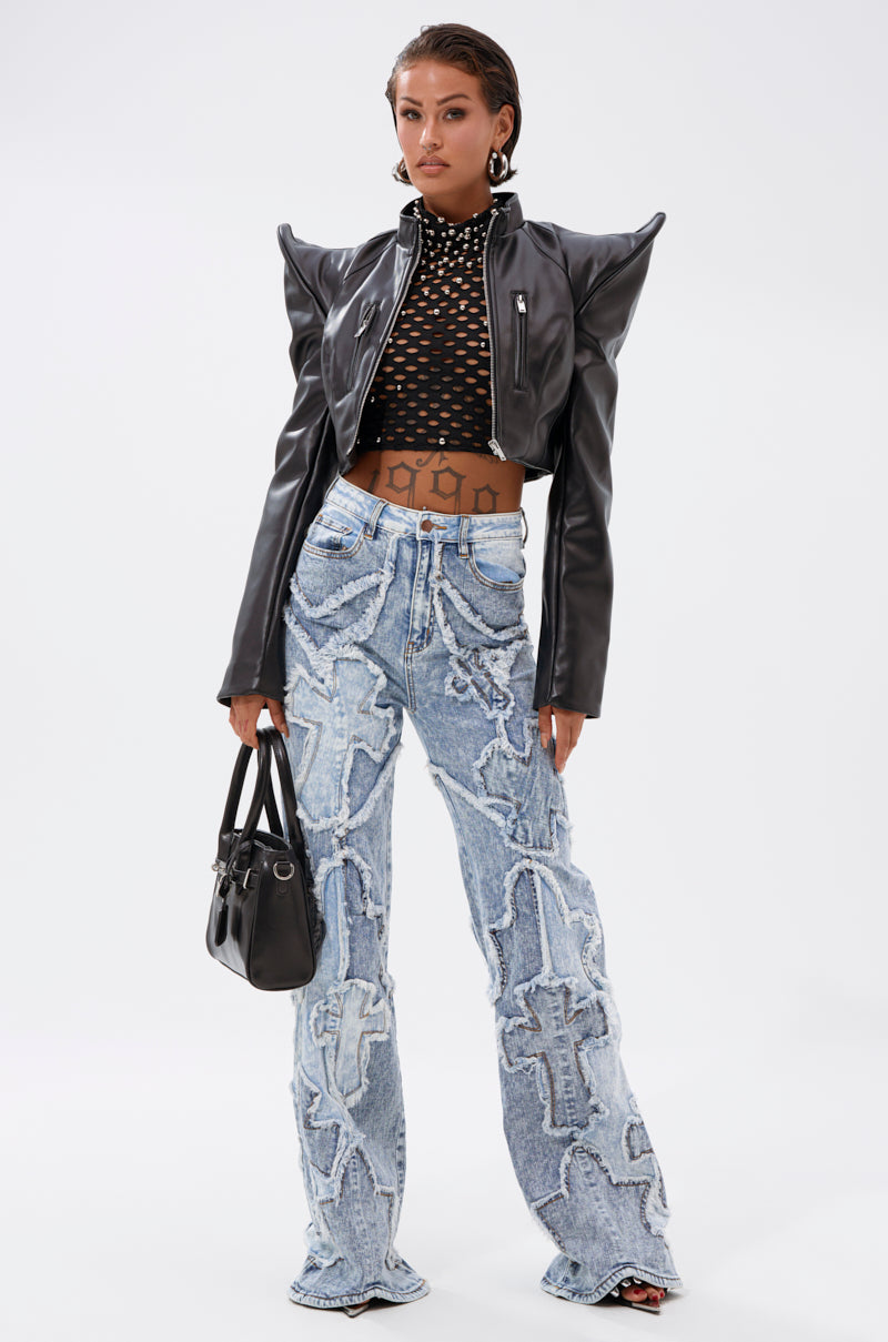 A woman stands confidently in a black leather jacket, black mesh crop top, and CROSSED OUT DENIM DISTRESSED PANT—a high-rise wide-leg jean with white appliqué—while holding a black handbag. 4