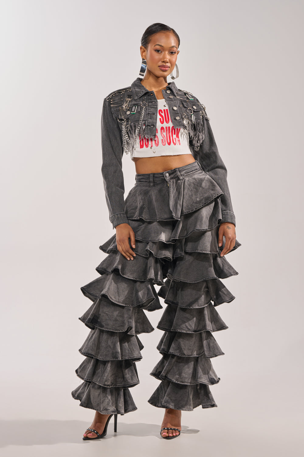 A woman stands confidently in the LOOK THIS WAY RUFFLE DENIM PANT, paired with a cropped graphic tee, a fringed studded denim jacket, and high heels against a plain light background. 0