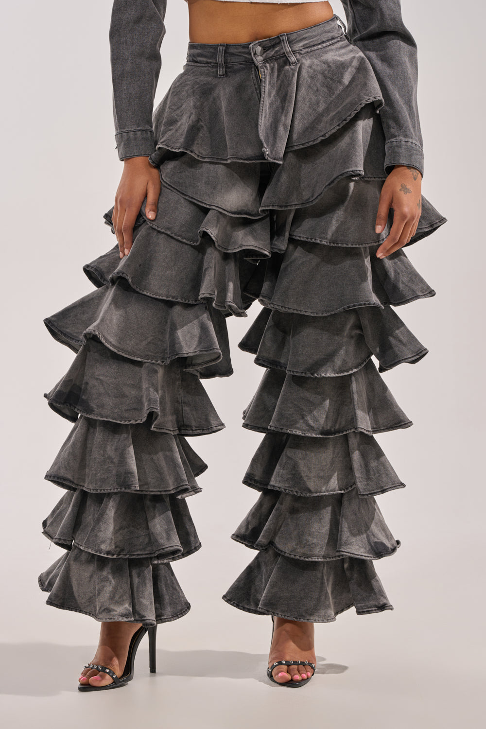 A person models the LOOK THIS WAY RUFFLE DENIM PANT—high-waisted, gray, flared jeans with dramatic layered ruffles—paired with black heeled sandals, shown from waist to feet against a plain background. 1