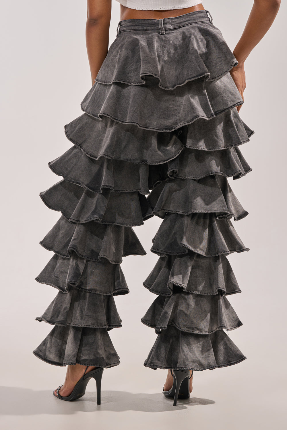 A person is shown from the back wearing the LOOK THIS WAY RUFFLE DENIM PANT—high-waisted, dark gray, wide-leg jeans with layered ruffles—paired with black high-heeled sandals against a plain background. 3
