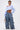 A person poses confidently in crop off-the-shoulder blue top and LOOK THIS WAY RUFFLE DENIM PANT with wide-leg silhouette, styled with white shoes, small patterned handbag, and short slicked-back hair.