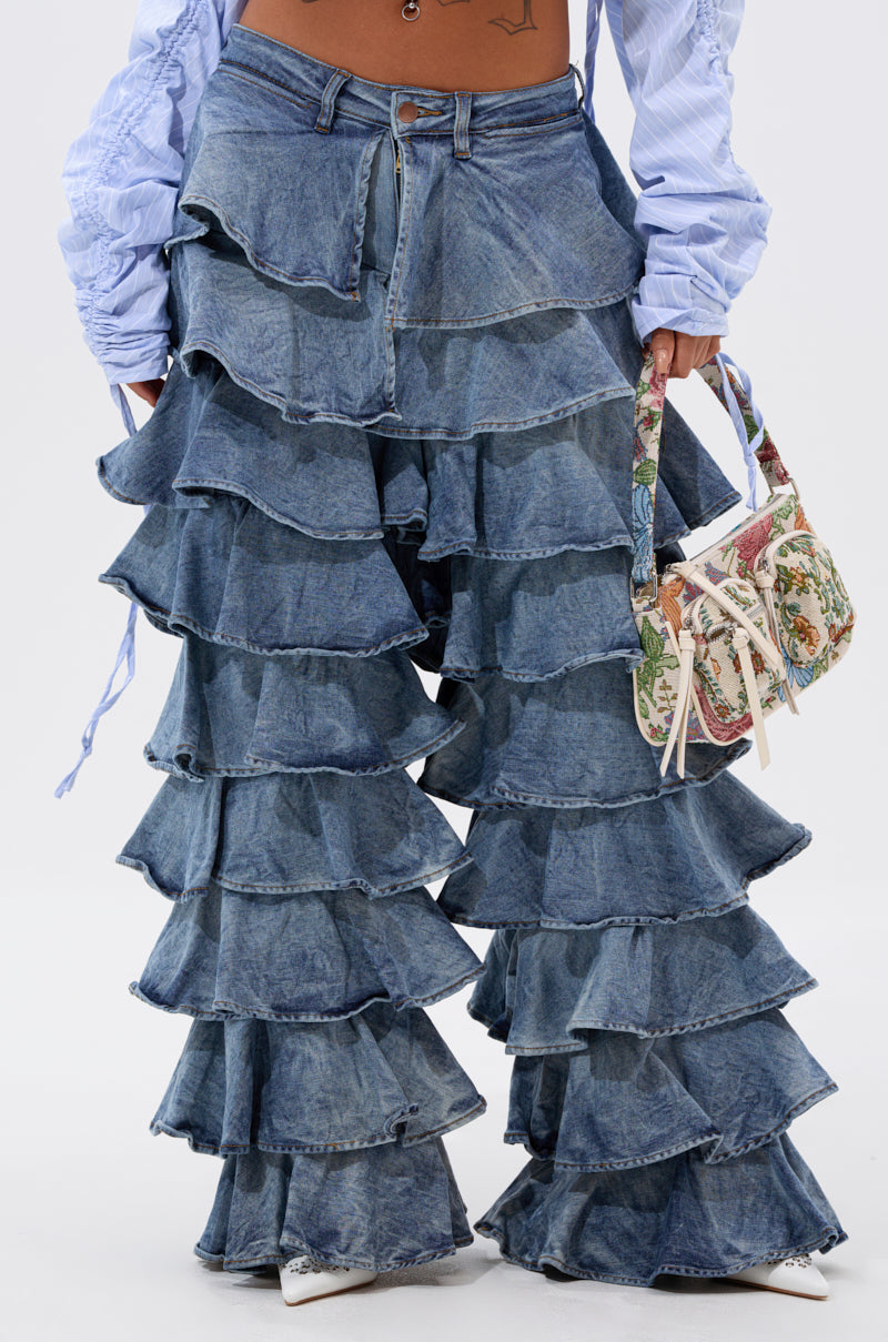 A cropped photo shows someone in LOOK THIS WAY RUFFLE DENIM PANT with multiple tiers, a tucked-in light blue shirt, white pointed shoes, and holding a small floral-patterned handbag.