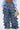 A cropped photo shows someone in LOOK THIS WAY RUFFLE DENIM PANT with multiple tiers, a tucked-in light blue shirt, white pointed shoes, and holding a small floral-patterned handbag.