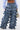 Wearing the LOOK THIS WAY RUFFLE DENIM PANT, a person shows off the high-rise, wide-leg ruffle style with a blue shirt and patterned handbag, their midriff visible against a plain light background.