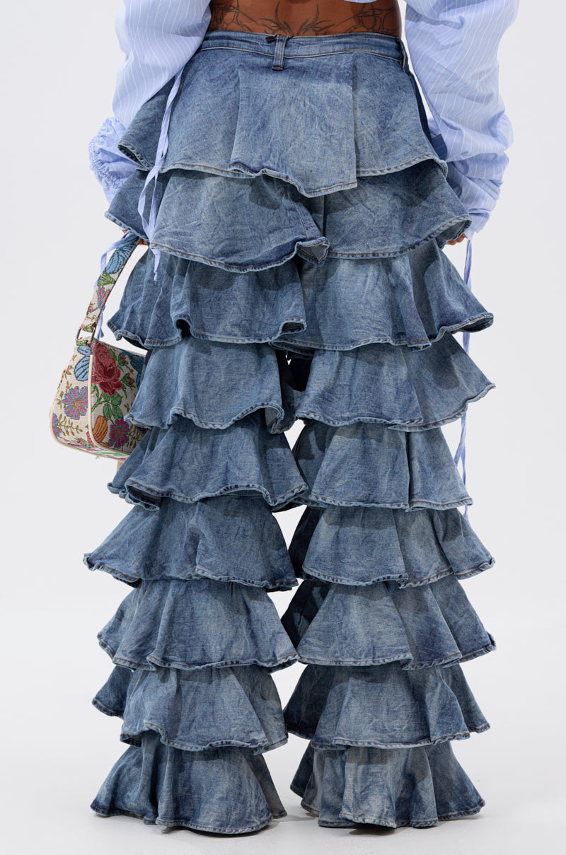 A person stands against a plain background, viewed from the back, wearing the LOOK THIS WAY RUFFLE DENIM PANT featuring high-rise multiple tiers and a wide-leg silhouette, styled with a blue shirt and floral handbag.