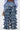 A person stands against a plain background, viewed from the back, wearing the LOOK THIS WAY RUFFLE DENIM PANT featuring high-rise multiple tiers and a wide-leg silhouette, styled with a blue shirt and floral handbag.