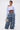 A woman poses in LOOK THIS WAY RUFFLE DENIM PANT paired with a light blue, off-the-shoulder crop top. She carries a patterned handbag, has short slicked-back hair, and reveals a 